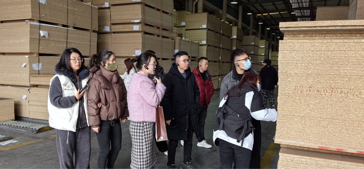 Visitors inspecting stacked raw material panels inside a factory warehouse.