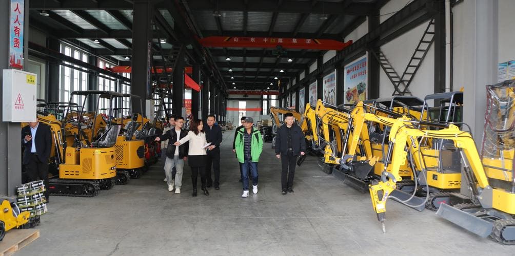 Buyers walking between rows of compact excavators in a machinery factory finished-goods hall.